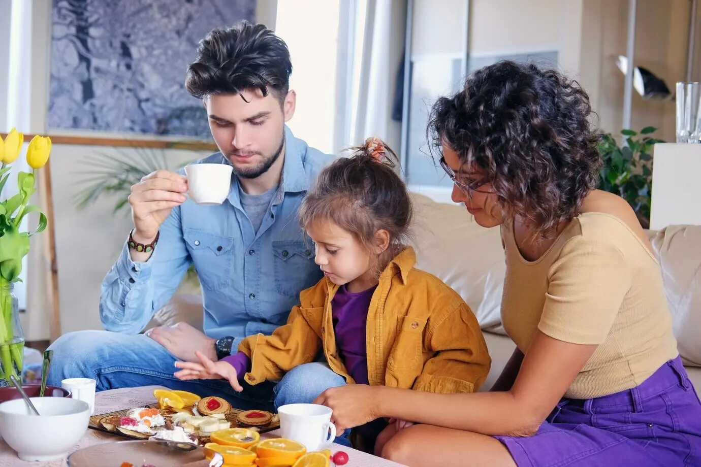 An attractive brunette woman wearing eyeglasses, a stylish bearded man, and their cute little daughter sit on a couch and eat lunch in a living room.